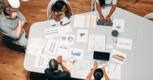 Top-down view of a meeting table with charts, graphs, papers, and laptops as four people discuss around it.