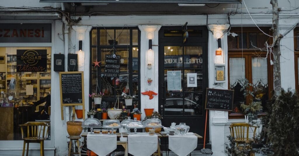 Outdoor cafe storefront with pastry displays under glass domes, chalkboard menus, and potted plants along a white facade storefront.