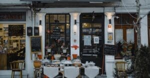 Outdoor cafe storefront with pastry displays under glass domes, chalkboard menus, and potted plants along a white facade storefront.