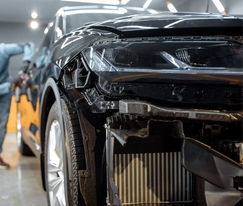 Front-left view of a black car with front-end damage in an auto repair shop, radiator exposed as repairs continue.