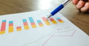 Close-up of a hand with a blue pen pointing to a multi-colored stacked bar chart on a printed sheet of paper.