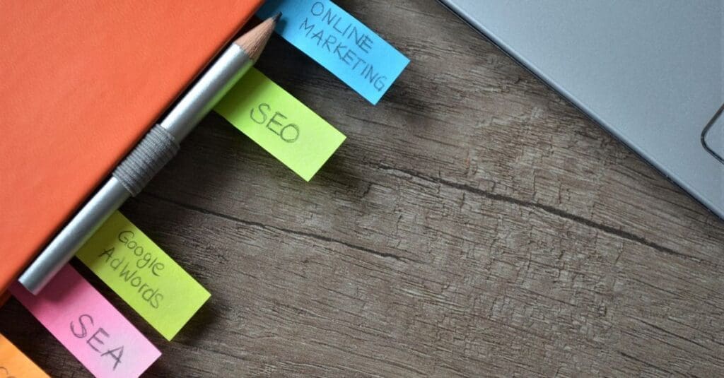 Top-down view of a workspace with an orange notebook, a pencil, and color‑coded sticky notes labeled Online Marketing, SEO, Google AdWords, and SEA near a laptop on a wooden desk.